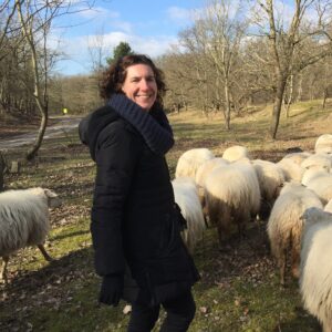 karin, eigenaar van buitendag, staat tussen de schapen in het duingebied, het is midden in de winter, de zon schijnt en een blauwe lucht is zichtbaar achter de bomen