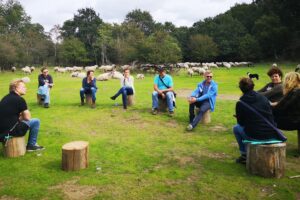 groep zit buiten op boomstammen naar elkaar te luisteren, ze zitten op een open plek met gras met bomen eromheen in duingebied met op de achtergrond kudde schapen